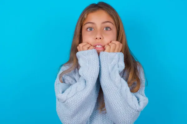 恐怖のブルネットの子供の女の子は青い背景に青いニットセーターを身に着けていることは 手を口の近くに保ち 恐怖と恐怖を感じ ショックと恐ろしい概念を持っています — ストック写真