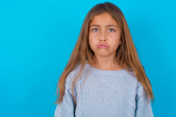 Displeased upset brunette kid girl wearing blue knitted sweater over blue background frowns face as going to cry, being discontent and unhappy as can\'t achieve goals,  Disappointed model has troubles