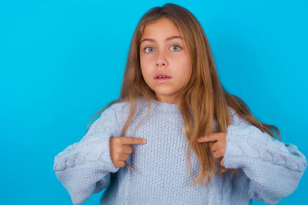 青い背景に青いニットセーターを着た恥ずかしいブルネットの子供の女の子は 困惑した表情で自分自身を示し 競争に参加することを選択されることにショックを受け 何かについて躊躇 — ストック写真