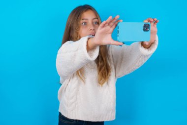 Esmer kız mavi arka plan üzerine beyaz örülü süveter giyip sosyal medyada yayınlamak için selfie çekiyor ya da arkadaşlarıyla görüntülü görüşme yapıyor..