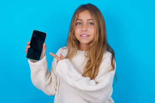 笑顔ブルネットの子供の女の子は青い背景の上に白いニットセーターを着てモックアップコピースペース 空白の画面を持つ携帯電話上のインデックス指を指す — ストック写真