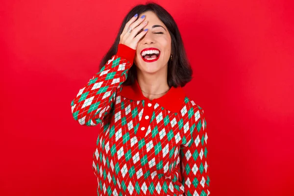 黑发女孩穿着针织毛衣 披着红色背景的圣诞礼服 满脸笑容 笑着肯定听到滑稽的笑话 — 图库照片