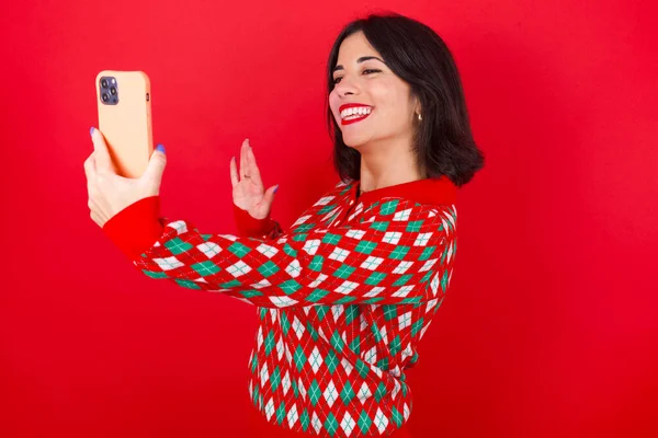 黑发高加索女孩身穿针织毛衣 背景为红色 手持现代手机 并在打招呼手势中做出视频呼叫波手掌 人民现代技术概念 — 图库照片