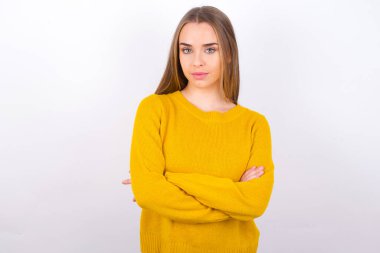 Stüdyoda kollarını kavuşturarak poz veren sarı kazaklı genç bir kadın.