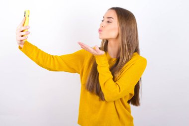 Stüdyoda akıllı telefonuyla poz veren genç bir kadın, kameraya öpücük yolluyor.