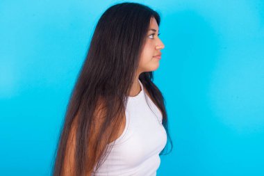 Mavi arka planda beyaz atlet giyen genç, mutlu, gülümseyen İspanyol kızın yan görüntüsü. 