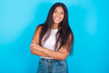 Mavi arka planda beyaz atlet giyen genç İspanyol kız gülümsüyor ve kameraya kendinden emin bir şekilde kollarını kavuşturuyordu. Pozitif ve kendinden emin kişi.