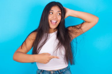 Mavi arka planda beyaz kolsuz bluz giyen genç İspanyol kız kendini olumlu hissediyor. Bu da bir şeyi gösteriyor. Bir elin başının üstünde, diğer elinle işaret et..