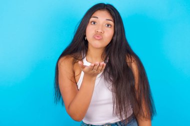 Mavi arka planda beyaz kolsuz bluz giyen genç İspanyol kız kameraya bakıp havada öpücükler savuruyor. Güzel ve seksi. Aşk ifadesi.