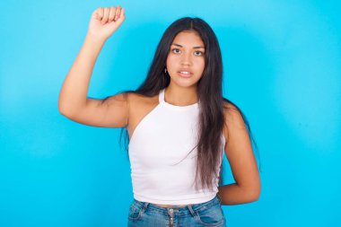 Mavi arka planda beyaz atlet giyen genç İspanyol kız ciddi, güçlü ve isyankar hissediyor, yumruğunu kaldırıyor, protesto ediyor veya devrim için savaşıyor..