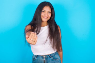 Mavi arka planda beyaz atlet giyen genç İspanyol kız, seni kendine güvenen ve gülümseyen, elinle hareket eden, pozitif ve arkadaş canlısı olmaya davet ediyor..