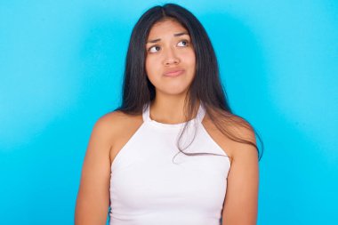 Mavi arka planda beyaz atlet giyen genç İspanyol kız, düşünceli bir şekilde kenara bakıyor, üniversiteden sonra ne yapacağını düşünüyor..