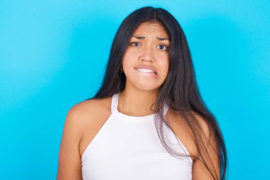 Mavi arka planda beyaz atlet giyen genç İspanyol kız gergin ve korkmuş dudakları ısıran sabırsız ifadeli kameraya bakıyor, düşünceli..