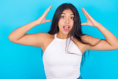 Mavi arka planda beyaz atlet giyen genç İspanyol kız kameraya heyecanla bakıyor, ellerini başının üstüne kaldırıyor beklenmedik bir şey fark ediyor, ani haberlere tepki veriyor..