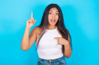 Mavi arka plan üzerine beyaz atlet giyen genç İspanyol kız diyor ki: Vay be, ne kadar heyecan verici, şaşırtıcı bir ifade, bir şey ifade ediyor. Bir eli göğsünde ve diğer eliyle işaret ediyor..