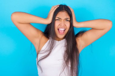 Paniğe kapılmış genç İspanyol kız mavi arka planda beyaz askılı bluz giyerek el ele tutuşup umutsuzluk ve hüsran içinde haykırıyor..