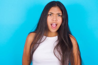 Etkileyici yüz ifadeleri. Şok olmuş genç İspanyol kız mavi arka planda beyaz kolsuz bluz giyiyor, çenesini düşük tutuyor gördüklerinden sersemlemiş hissediyor.