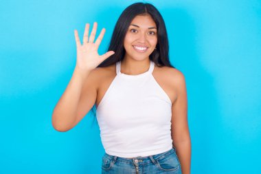 Mavi arka planda beyaz atlet giyen genç İspanyol kız beş numaralı parmaklarıyla gülümseyerek kendinden emin ve mutlu olduğunu gösteriyor..