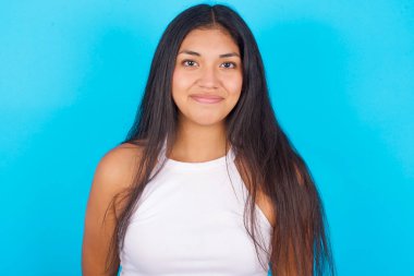 Mavi arka planda beyaz atlet giyen mutlu, genç İspanyol kız büyüleyici bir gülümsemeyle kameraya bakıyor..