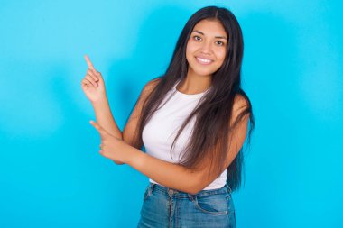 Mavi arkaplan üzerine beyaz atlet giyen genç İspanyol kız, tanıtım metnin ya da ilanın için boş fotokopi alanında ön parmağınla işaret ediyor..
