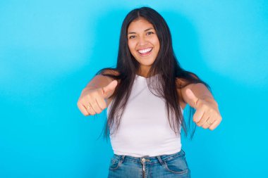 Mavi arka planda beyaz kolsuz bluz giyen genç İspanyol kız olumlu bir jest yapıyor. Başparmağıyla gülümsüyor ve başarı için seviniyor. Kameraya bak, kazanan jest..