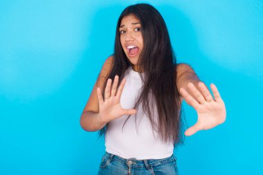 Tatminsiz genç İspanyol kız beyaz kolsuz bluz giyiyor mavi arka planda kaşlarını çatıyor, iğrenç bir ifade takınıyor, dilini gösteriyor, itaatsizlik ediyor, birine sinir oluyor..