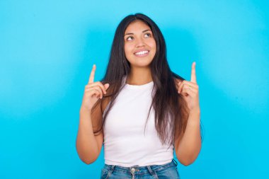 Başarılı, dost canlısı görünümlü, beyaz kolsuz bluz giyen, mavi arka planda heyecanla bağıran, her iki işaret parmağını kaldıran, bir şeyi işaret eden genç İspanyol kız..