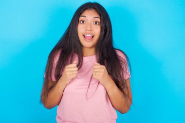 Mavi arka planda pembe tişört giyen çaresiz ve şok olmuş genç bir kızın portresi paniğe kapılmış görünüyor, el ele tutuşuyor, ağzı açık..