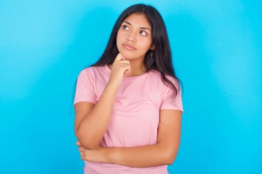 Yüz ifadeleri ve duygular. Mavi arka plan üzerine pembe tişört giyen düşünceli, genç İspanyol kız başının altında el ele tutuşuyor..