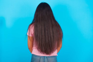 Mavi arka planda pembe tişört giyen genç İspanyol kız kolları vücudunda geriye doğru bakıyor..