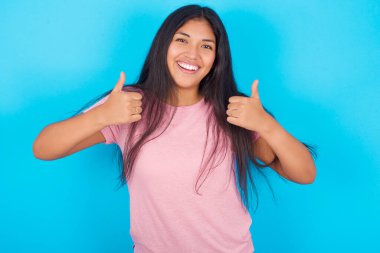 İyimser genç İspanyol kız pembe tişört giyip mavi arka plana olumlu duygular besliyor. Kalite ve tavsiye kavramı.