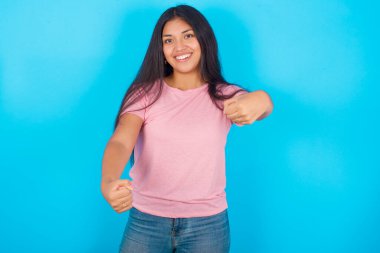 Mavi arka planda pembe tişört giyen, ellerini başının üstünde tutarak gülümseyen çekici İspanyol kızın portresi. Güven ve rahatlama kavramı.