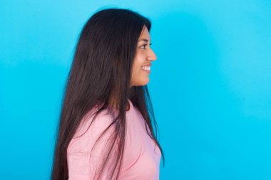 Gülümseyen İspanyol kızın profili pembe tişörtlü, mavi arkaplanlı, sağlıklı ciltli, derin düşüncelere dalmış, açık havada yürümeye hazır..