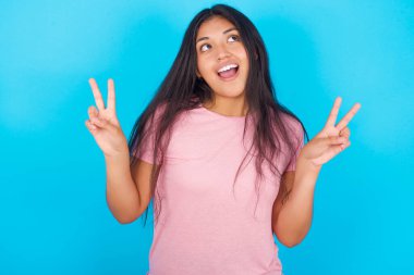 Mavi arka planda pembe tişört giyen neşeli, genç bir kızın tek pozu iki elinle barış ya da zafer işareti yapar..