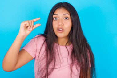 Mavi arka planda pembe tişört giyen, şoke olmuş genç İspanyol kız elleri olan, boyunu gösteren, ağzında şaşkınlığı gösteren bir şey gösteriyor. Ölçüm kavramı.
