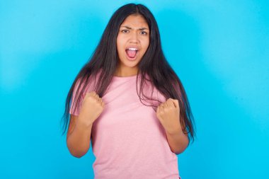 Neşeli, heyecanlı, şanslı, pembe tişörtlü İspanyol kız mavi arka planda tezahürat yapıyor, başarıyı kutluyor, yumruklarını sıkarak evet diye bağırıyor.