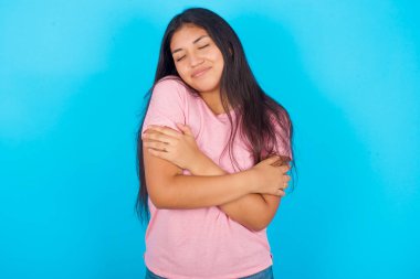 MODEL. Mutlu ve olumlu bir şekilde kendine sarılmak, kendinden emin gülümsemek. Özgüven ve özgüven. Mavi arka planda pembe tişört giyen İspanyol genç kız.