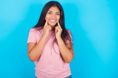 Güçlü, sağlıklı, beyaz dişler. Mavi arka planda pembe tişört giyen mutlu İspanyol kızın portresi. Parlayan gülümsemesi kusursuz beyaz dişleri işaret ediyor..