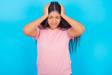 Mavi arka planda pembe tişört giyen genç İspanyol kız elleriyle başını tutuyor şiddetli baş ağrısından muzdarip, parmaklarını şakaklarına bastırıyor.