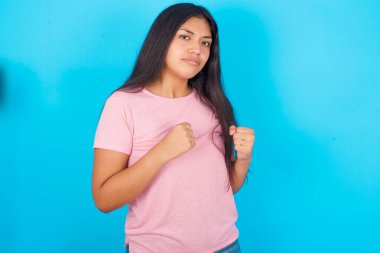 Çekici İspanyol kızın portresi. Mavi arka plan üzerine pembe tişört giymiş. Boks pozisyonunda önünde el ele tutuşmuş..