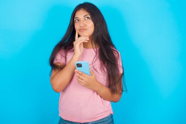 Mavi arka planda pembe tişört giyen İspanyol bir kızın cep telefonu kullandığını ve el ele tutuştuğunu hayal ediyorum. Karar almak ve sosyal medya kavramı.