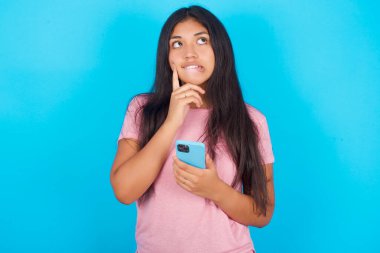 Düşünen genç bir İspanyol kızın mavi arka planda pembe tişört giyip cep telefonunu kullanırken ve el ele tutuşurken görüntüsü. Karar almak ve sosyal medya kavramı.