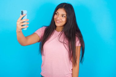 Mavi arka planda pembe tişört giyen mutlu, genç İspanyol bir kızın tek pozu cep telefonuyla selfie çekiyor. İnsanlar, teknoloji ve eğlence kavramı