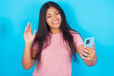 Mutlu, dost canlısı İspanyol kızın portresi pembe tişört giymiş mavi arka planda selfie çekiyor ve el sallıyor, video görüşmesinde iletişim kuruyor, internette sohbet ediyor..