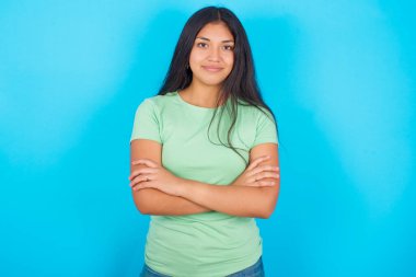 Kendine güveni tam, sakin, mavi arka planda yeşil tişört giyen İspanyol kız kollarını kavuşturmuş duruyor. Profesyonel titreşimlerin kendine güvenen pozlarda durduğunu gösterir.