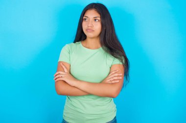 Mavi arka planda yeşil tişört giyen düşünceli, düşünceli, genç bir İspanyol kız kollarını kavuşturmuş, düşünceli bir ifadeyle ne yapacağını düşünüyor.