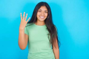 Mavi arka planda yeşil tişört giyen genç İspanyol kız gülümsüyor ve arkadaş canlısı görünüyor, dört ya da dört numarayı gösteriyor, geriye doğru sayıyor.