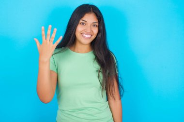 Mavi arka planda yeşil tişört giyen genç İspanyol kız gülümsüyor ve arkadaş canlısı görünüyor, 5 ya da 5 numarayı gösteriyor, geri sayıyor.