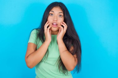 Dilsiz, mavi arka planda yeşil tişört giyen genç İspanyol kız kameraya şoke edici bakışlara karşı elleri açık, ağzı açık tepki veriyor.