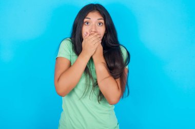 Sersemlemiş genç İspanyol kız mavi arka planda yeşil tişört giyiyor iki eliyle ağzını kapatıyor çarpıcı dedikodular duyduktan sonra ya da bir şeyden korkarak..